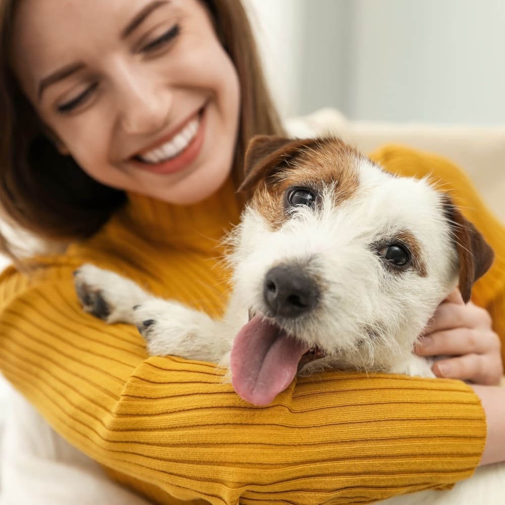 Woman in a yellow sweater smiling and holding a small white dog who has their mouth open and tongue hanging out